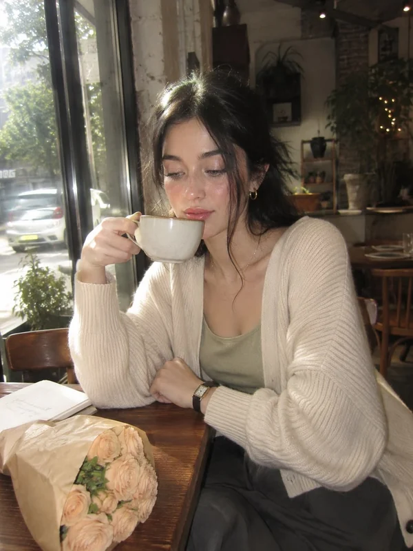 Cream cardigan and sage top, cup near lips at a sunlit cafe window with bouquet